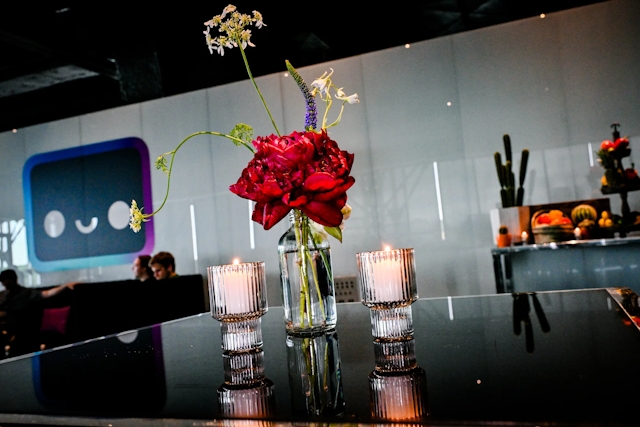 A rose floral in a small vase with water in between two candles on a high black table. In the background is a taco bar to the right and a lounge with a large Houston decal to the left.