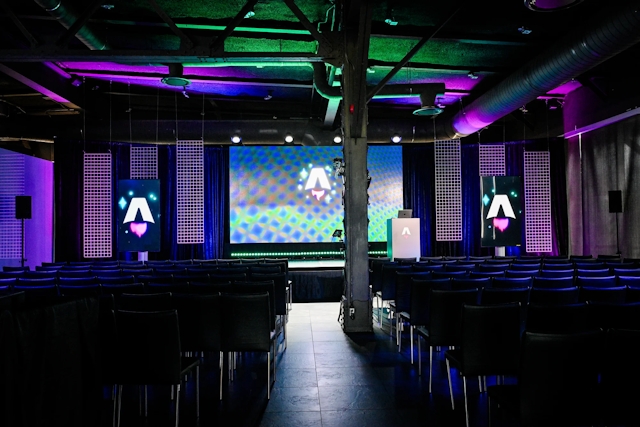 View from the back of the dark empty keynote presentation area. Rows empty black chairs. A vibrant large LED screen with the Astro Together logo on it. Two smaller portrait-oriented screens adorn the sides of the stage, also with the Astro Together logo.