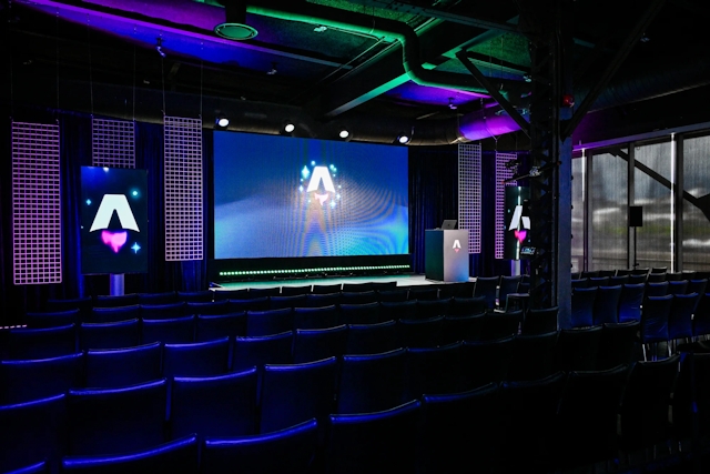 View from the back of the dark empty keynote presentation area. Rows empty black chairs. A vibrant large LED screen with the Astro Together logo on it. Two smaller portrait-oriented screens adorn the sides of the stage, also with the Astro Together logo.