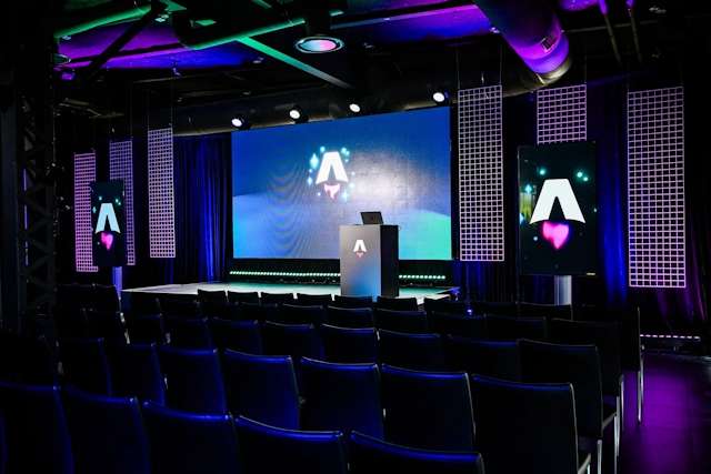 View from the back of the dark empty keynote presentation area. Rows empty black chairs. A vibrant large LED screen with the Astro Together logo on it. Two smaller portrait-oriented screens adorn the sides of the stage, also with the Astro Together logo.