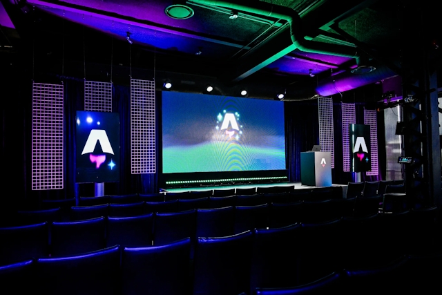 View from the back of the dark empty keynote presentation area. Rows empty black chairs. A vibrant large LED screen with the Astro Together logo on it. Two smaller portrait-oriented screens adorn the sides of the stage, also with the Astro Together logo.