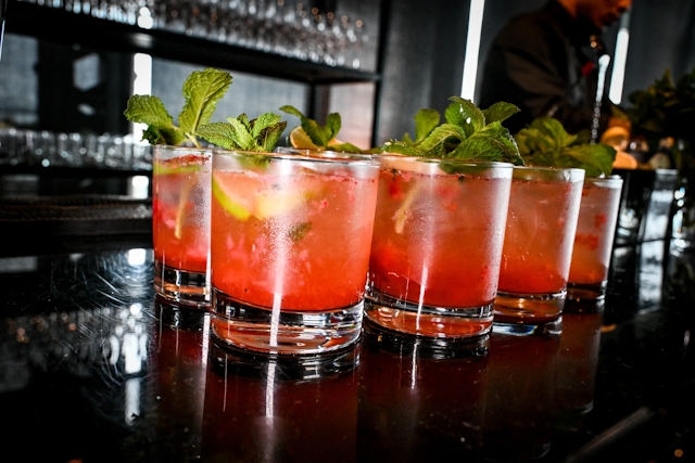 Half a dozen pink cocktails garnished with mint and lemon atop a black bar.