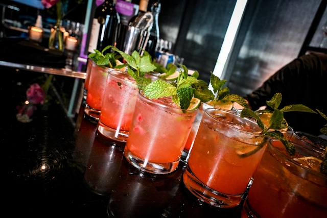 Half a dozen pink cocktails garnished with mint and lemon atop a black bar.