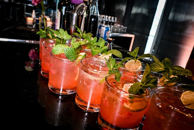 Half a dozen pink cocktails garnished with mint and lemon atop a black bar.