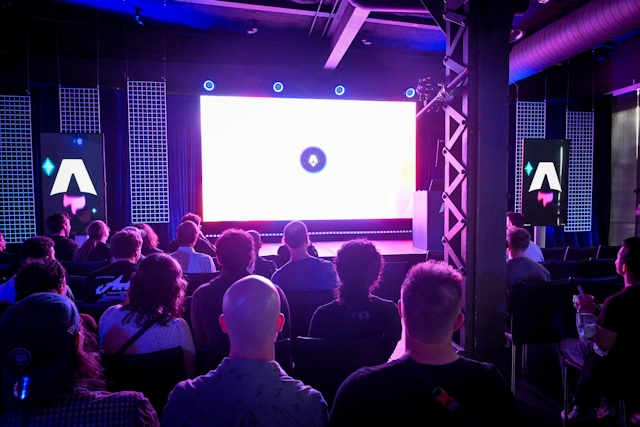 View from the back of guests in their seats, waiting for the keynote presentation to begin. They are brightly lit in purple by the large LED screen on stage.