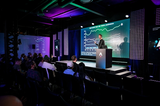 Fred K. Schott on stage in front of a slide depicting a graph, standing next a podium with a laptop sitting atop it. Wide shot including the audience.