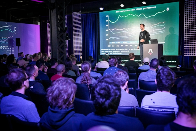 View from the back of the audience. Fred K. Schott is on stage with a slide depicting a graph, standing next to a branded Astro podium with a laptop on top of it.