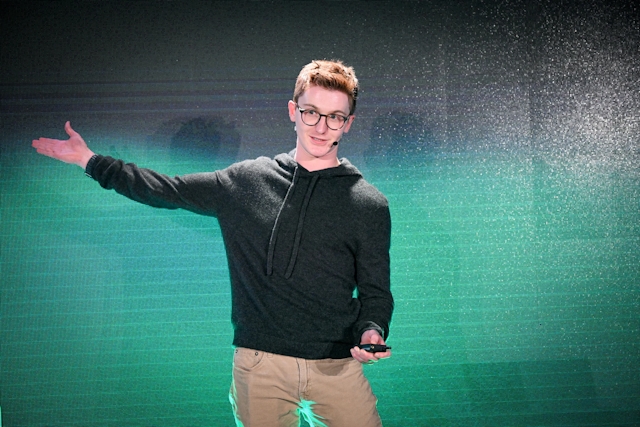 Ben Holmes on stage in front of a green and black LED screen. His right arm is outstretched, pointing to something off-camera.