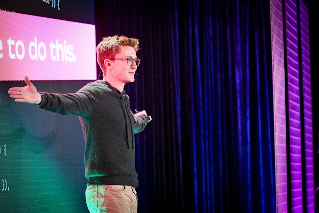 A close up of Ben Holmes on stage with both arms outstretched, gazing at the audience. He is lit with pink and purple lighting.