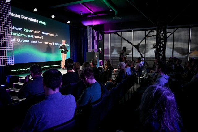 A shot from the side of the audience. Ben Holmes stands on stage in front of a code snippet depicting a form. The slide reads, "Make FormData easy."