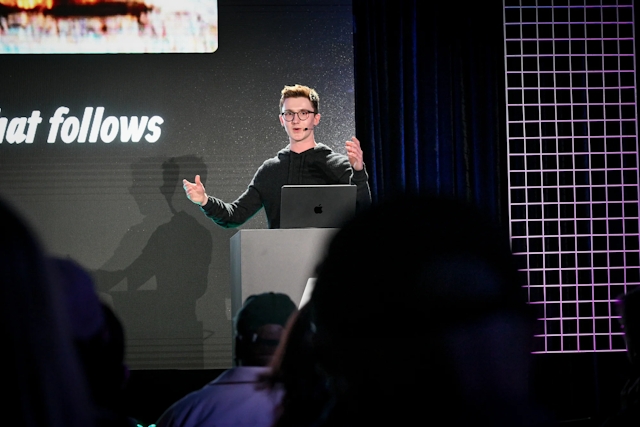 Ben Holmes speaking from an Astro-branded podium. Both his hands are up as he addresses the audience.