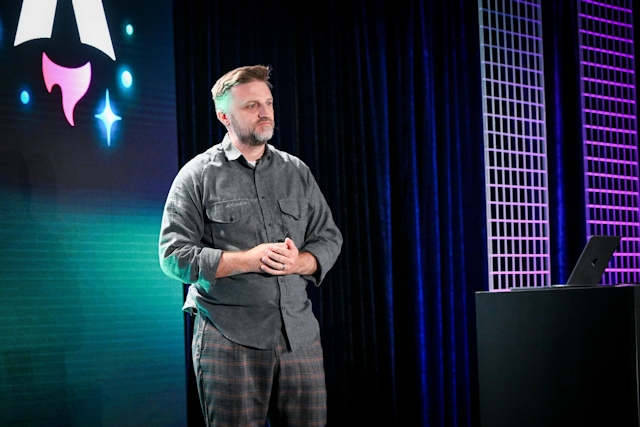 Matthew Phillips speaking on stage next to a podium with a laptop on it. Behind him is the Astro Together logo on-screen.