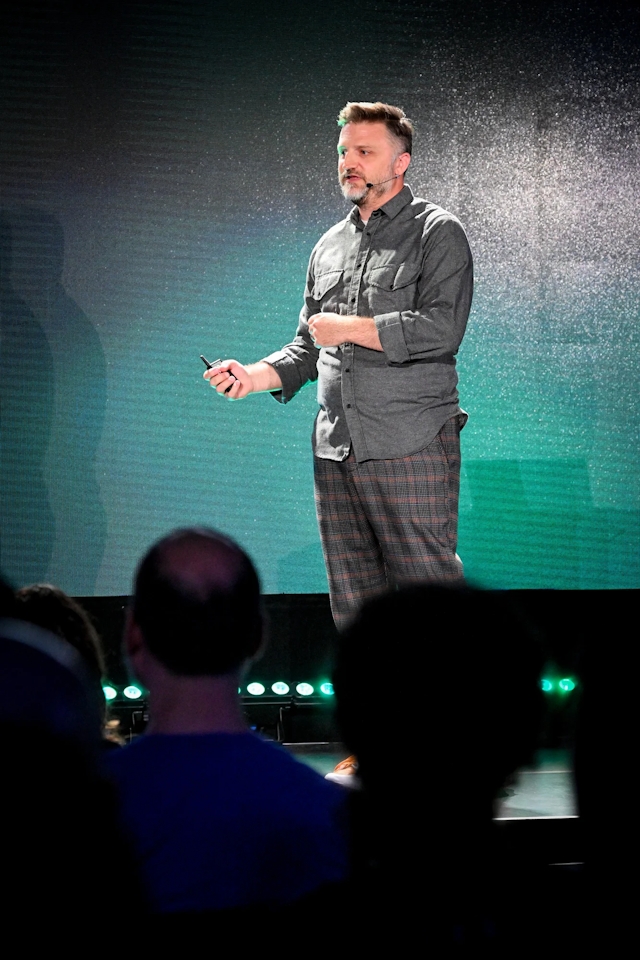 Portrait view of Matthew Phillips speaking on stage with audience members in the foreground.