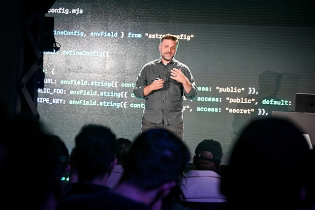 View from the audience of Matthew Phillips speaking on stage, standing in front of a code example.