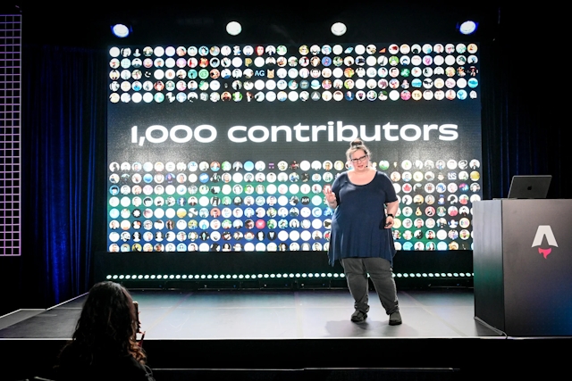 Sarah Rainsberger speaking from the right side of the stage, talking about how Docs has had 1,000 contributors to-date.