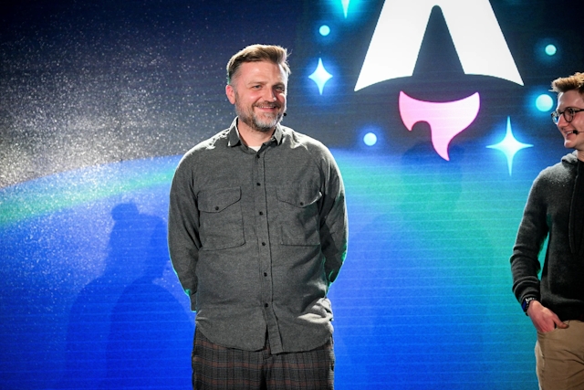 Closeup of Matthew Phillips standing with other keynote speakers on stage. His hands are behind his back and he is smiling at the audience. Off to the right, Ben Holmes is smiling at him.