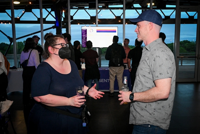 Sarah Rainsberger, wearing a black mask, chats with a guest. The Sentry booth and TV screen can be seen behind them.